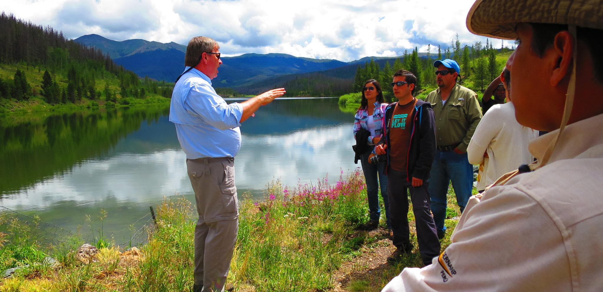 man talking to a group of people by a mountain lake