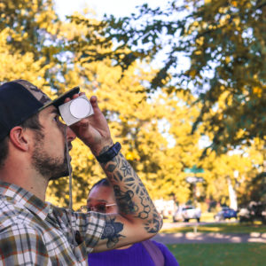 A sustainability master's student looks through a rangefinder in a park, surrounded by trees.