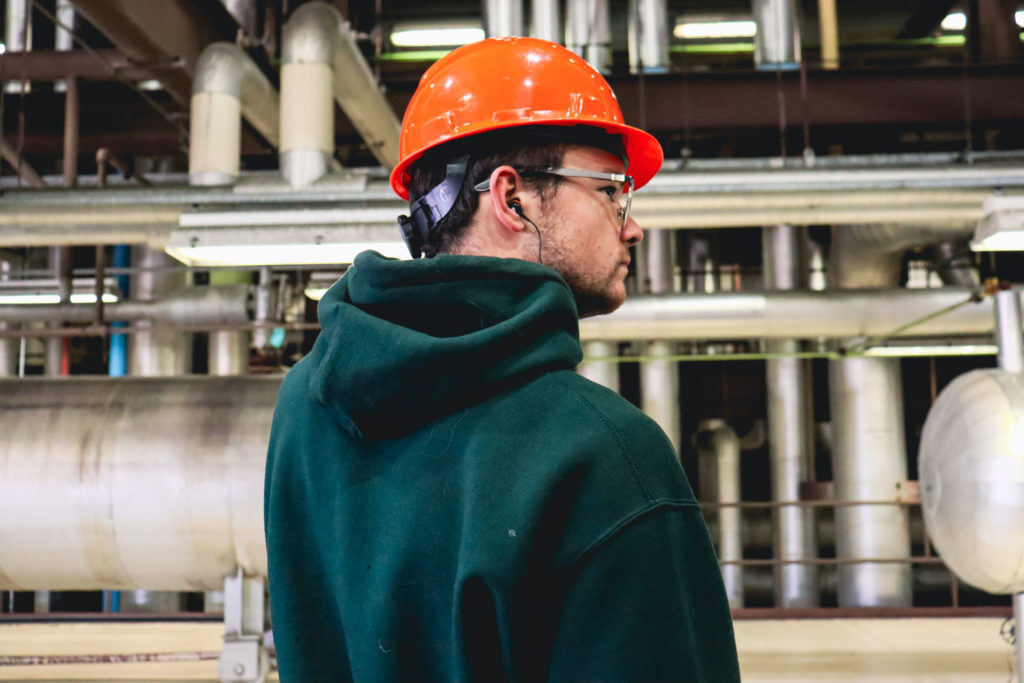 Student in green sweatshirt and orange hardhat standing in front of many metal pipes in an energy plant.