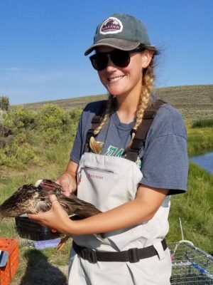Casey wearing outdoor gear and sunglasses smiles while holding a duck in her hands. She stands in a grassy area near a pond under a clear blue sky.