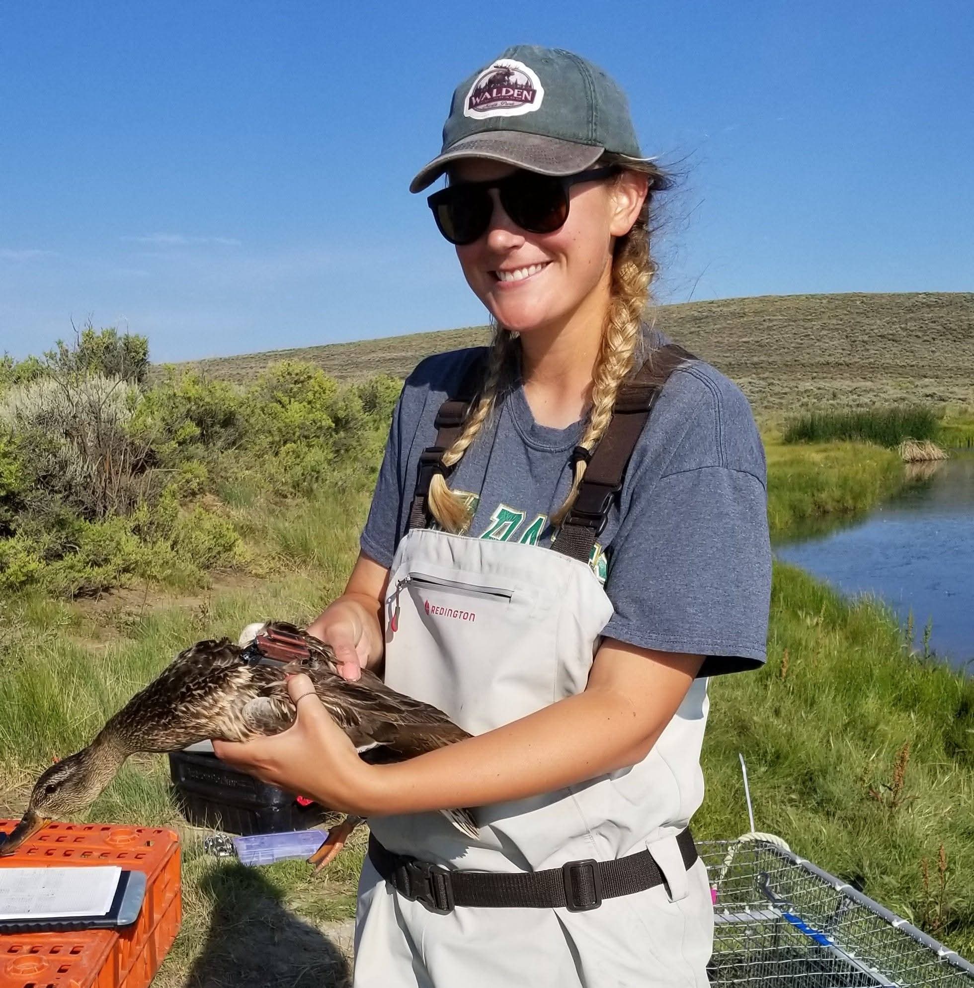 Casey wearing outdoor gear and sunglasses smiles while holding a duck in her hands. She stands in a grassy area near a pond under a clear blue sky.