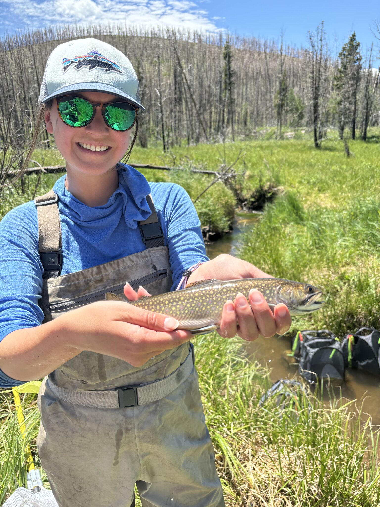 Kim, wearing outdoor gear and sunglasses smiles while holding a fish in her hands by a stream in a lush, green meadow.