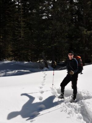 Person hiking through deep snow in a forested area, wearing winter clothing, sunglasses, and a backpack, and using trekking poles; footprints trail behind them in the snow.